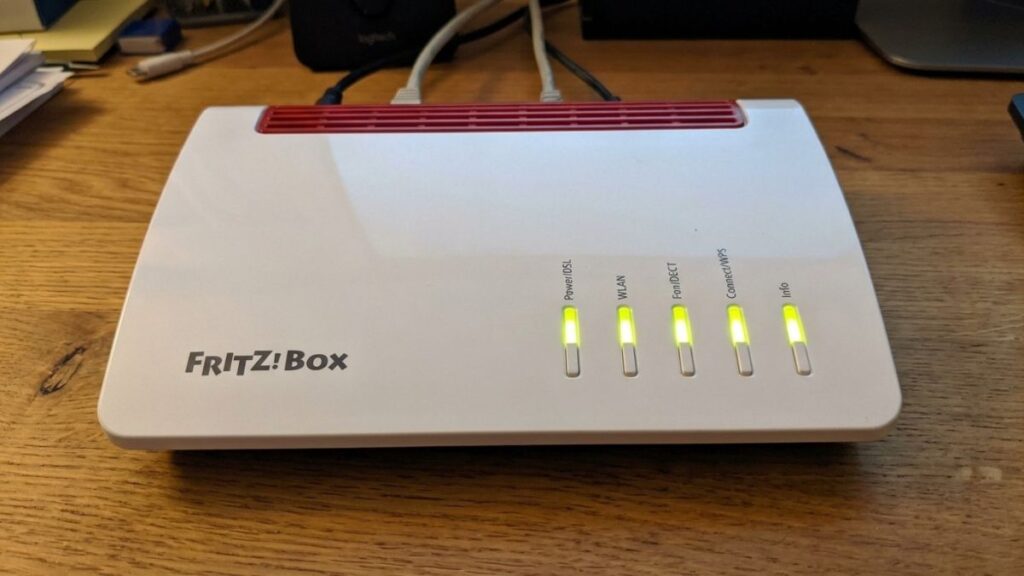 Top-down view of a white and red FRITZ!Box router on a wooden table, with all five LED status lights glowing solid green for Power/DSL, WLAN, Fon/DECT, Connect/WPS, and Info.