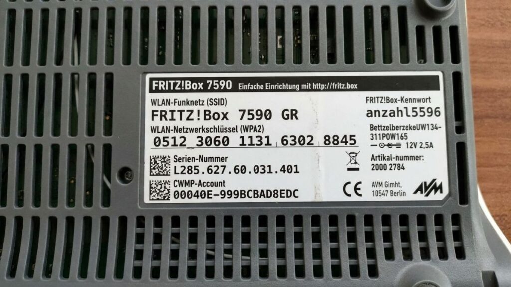 A close-up photograph of the product label sticker on the underside of a grey FRITZ!Box 7590 router, detailing the default login URL [http://fritz.box](http://fritz.box), SSID FRITZ!Box 7590 GR, and the FRITZ!Box-Kennwort admin password.