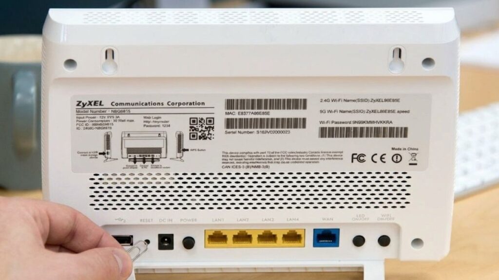 A hand using a straightened paperclip to press the recessed RESET button on the back panel of a white Zyxel NBG6815 router, next to the DC IN power port and LAN ports.